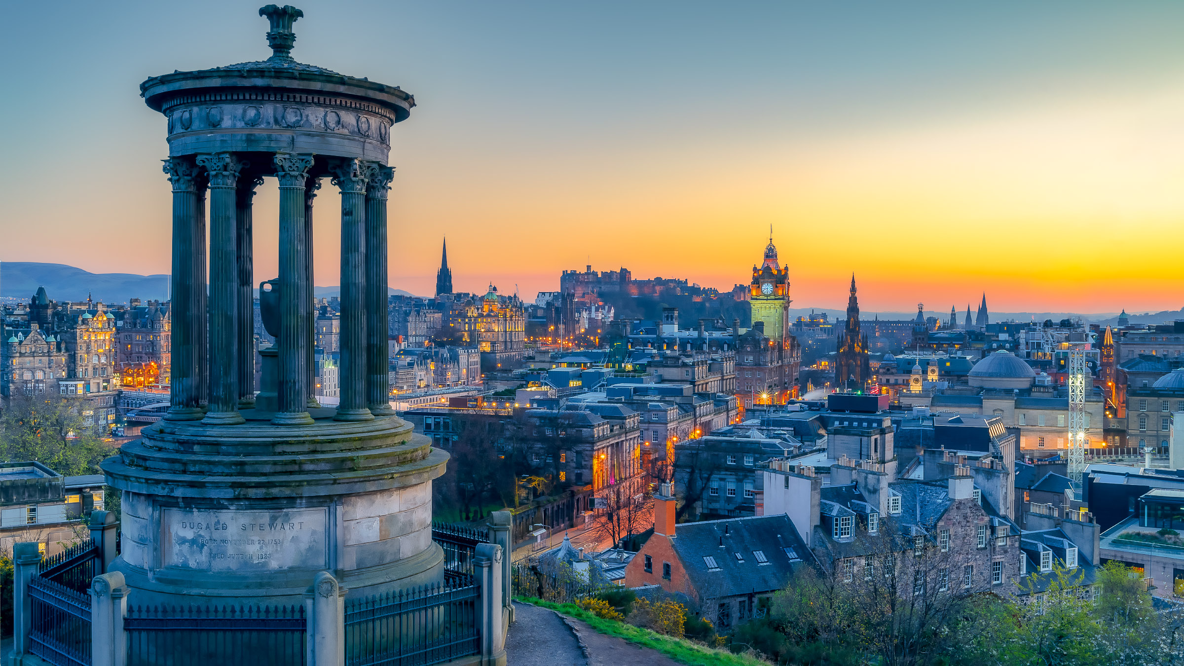 Edinburgh skyline at sunset from Calton Hill, the Dugald Stewart Monument in the foreground before the lit Old Town.